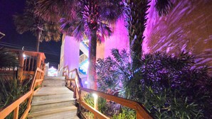 A wooden staircase with railings, surrounded by tropical plants and palm trees, leading to a building with illuminated purple lighting.