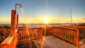 A wooden deck with a showerhead, overlooking a beach and a pier during sunset.