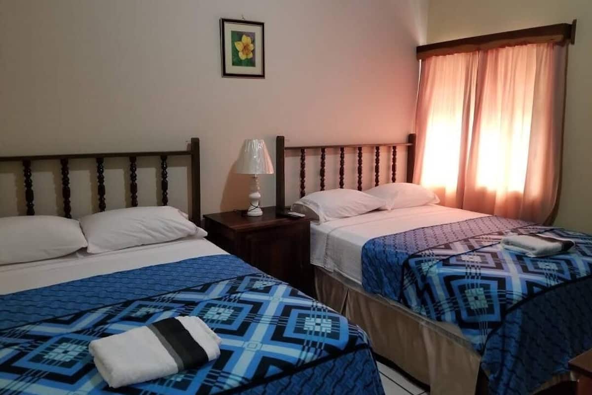 Habitación de hotel con dos camas, una mesita de noche con lámpara y una ventana con cortinas.