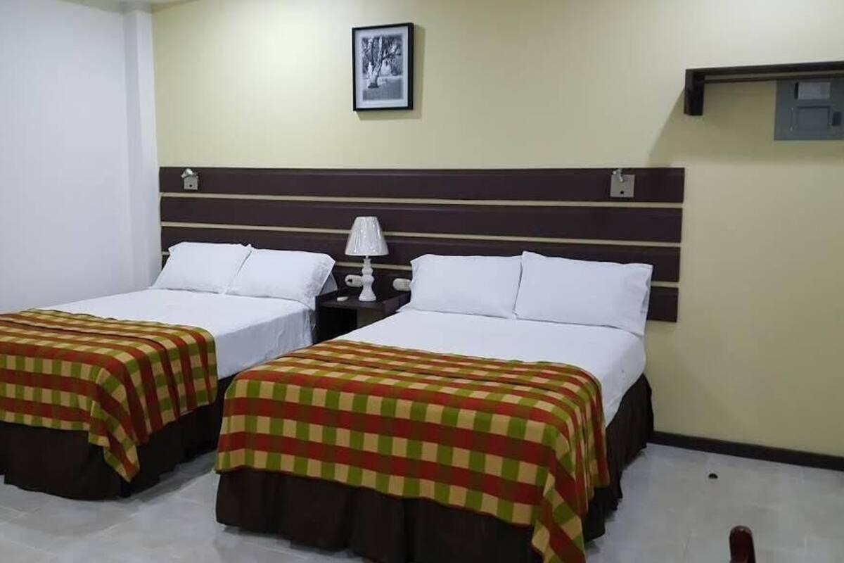 Habitación de hotel con dos camas individuales, cada una con colcha a cuadros y almohadas blancas. Entre las camas hay una mesita de noche con una lámpara, y sobre ellas, un cuadro en la pared.
