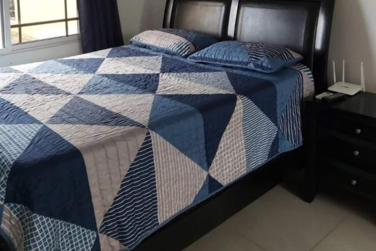Un dormitorio con cama, ventana con cortinas, un cuadro colgado en la pared y una mesita de noche con lámpara.