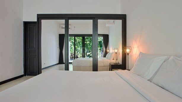 A bedroom with a large bed, a nightstand with a lamp, and a view of a garden through a sliding door. - Villa Arus (Seminyak)