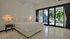 A bedroom with a bed, two bedside lamps, a window with a view of greenery, and a ceiling air conditioner. - Villa Arus (Seminyak)