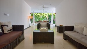 A modern living room with wicker furniture, a glass coffee table, and a view of lush greenery outside. - Villa Arus (Seminyak)