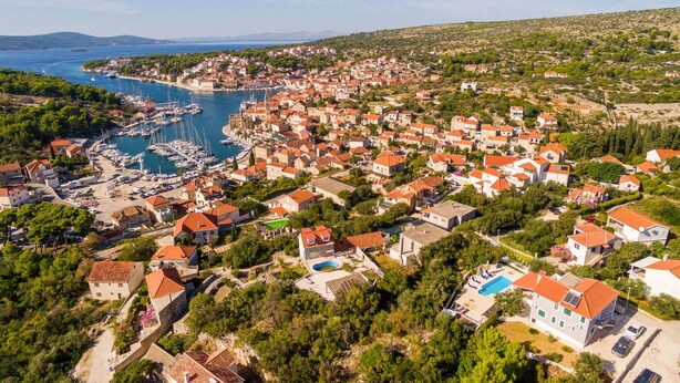 Un pueblo costero con un puerto deportivo, casas con tejados rojos y un paisaje montañoso de fondo.