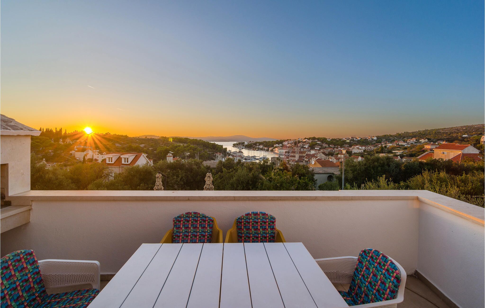 A balcony with a white table and chairs, overlooking a coastal town during sunset.