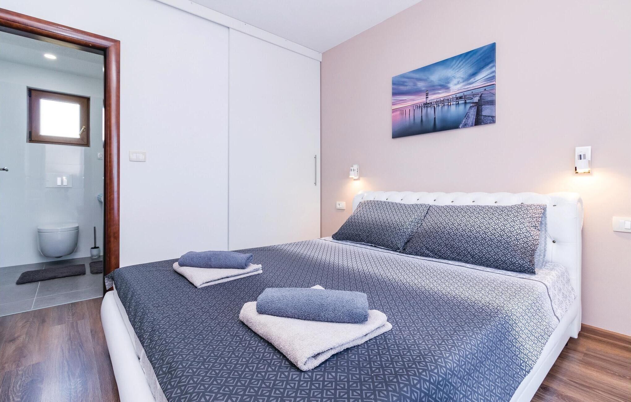 A bedroom with a bed, pillows, a painting, and a bathroom visible through an open door.