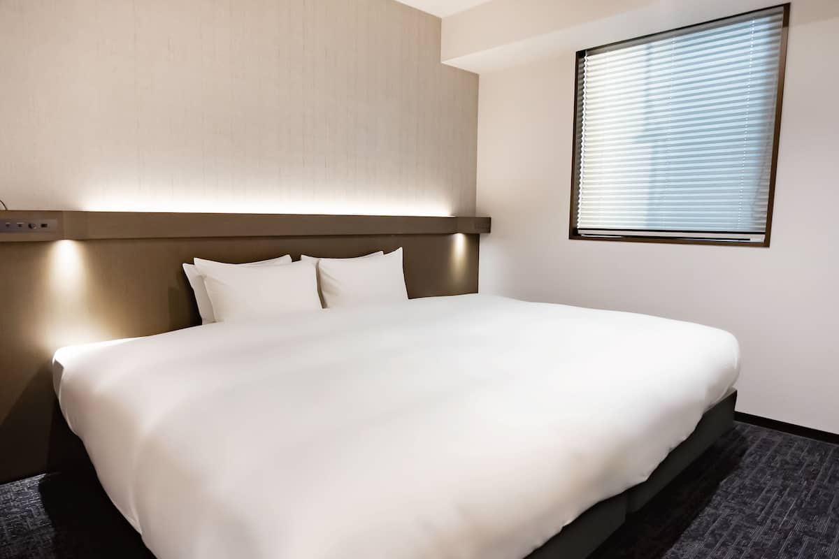 A neatly made bed in a hotel room with a headboard, a control panel, and a window with blinds.