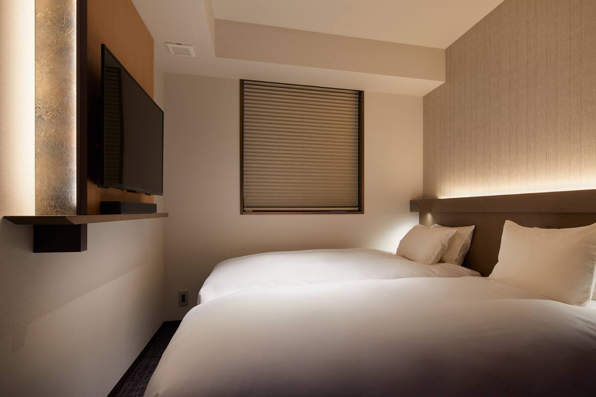 A modern hotel room with a large bed, a flat-screen TV mounted on the wall, and a window with blinds.