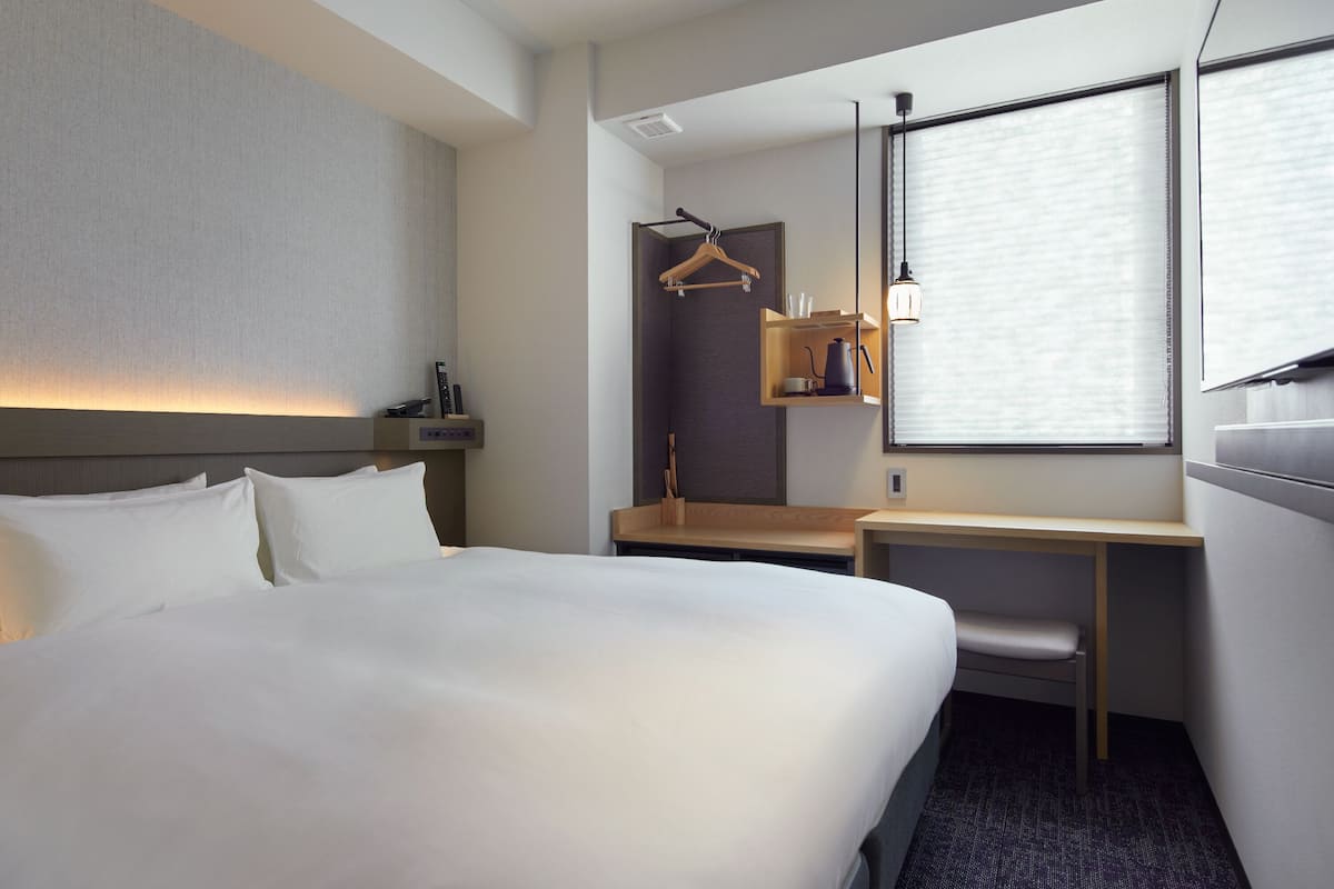 A hotel room with a large bed, a desk, a chair, a wardrobe, and a window with blinds.