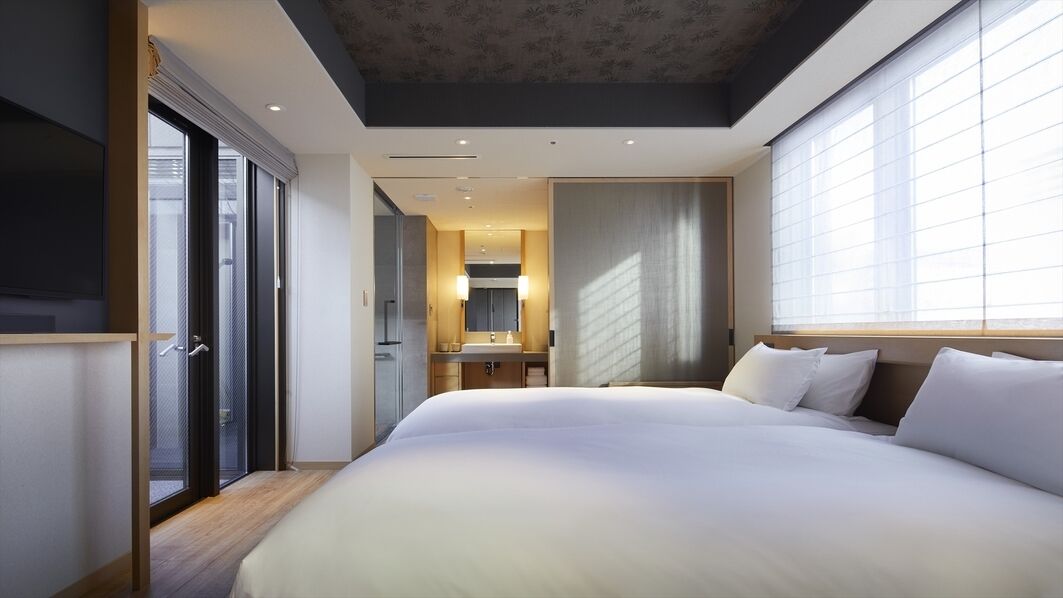 A modern hotel room with a large bed, a bathroom with a mirror, and a view of the city through large windows.