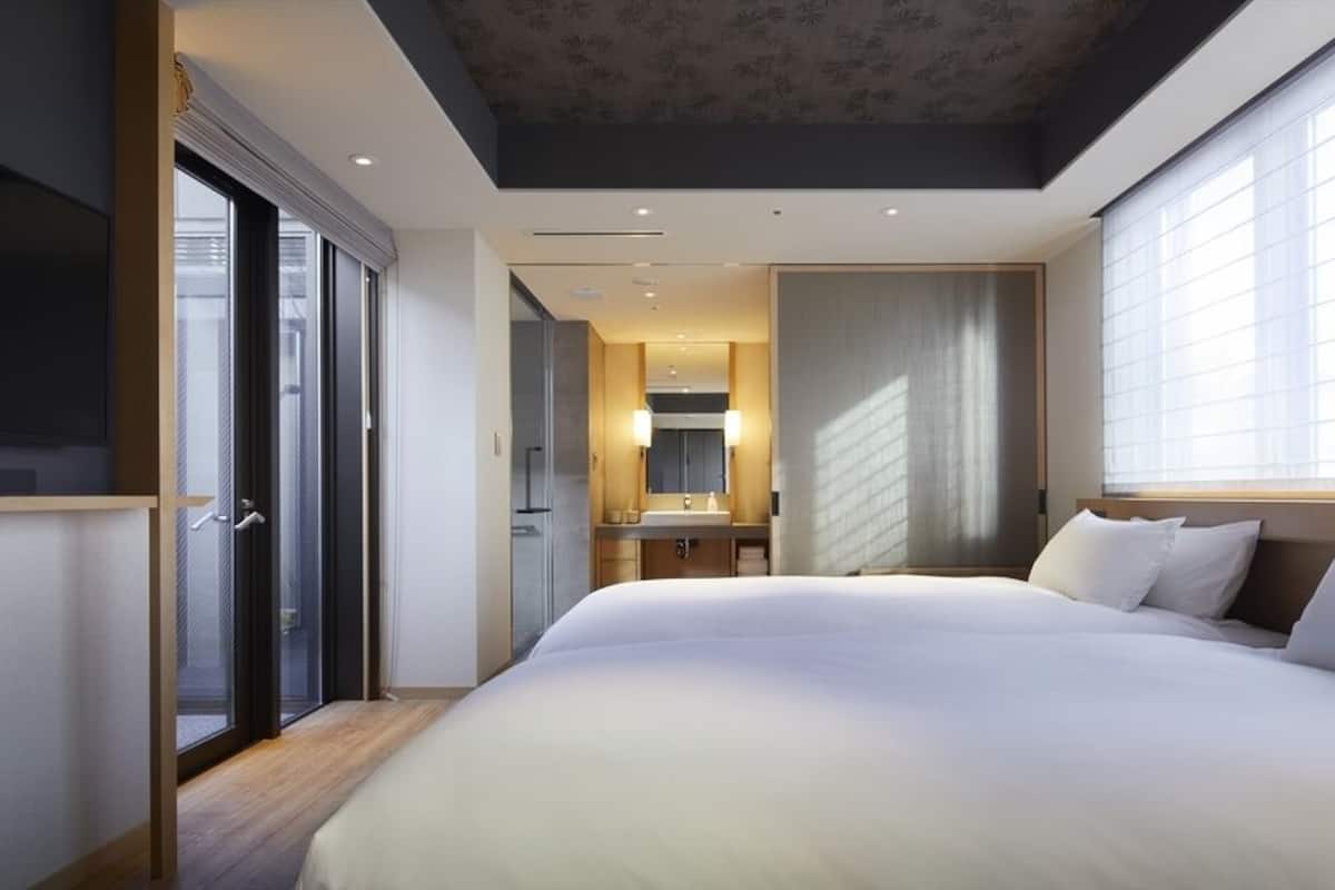 A modern hotel room with a large bed, a bathroom with a mirror, and a view of the city through large windows.
