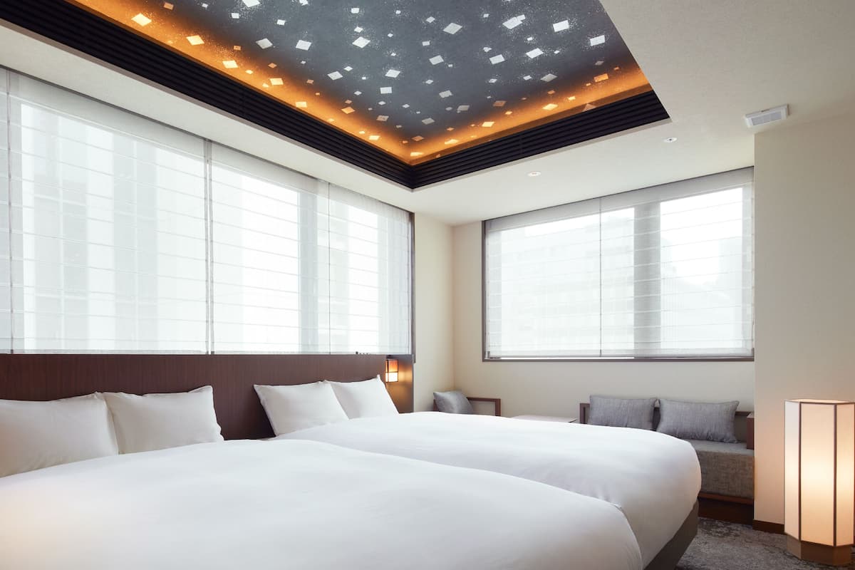 A hotel room with two large beds, a ceiling with recessed lighting, large windows, and a seating area with a lamp.