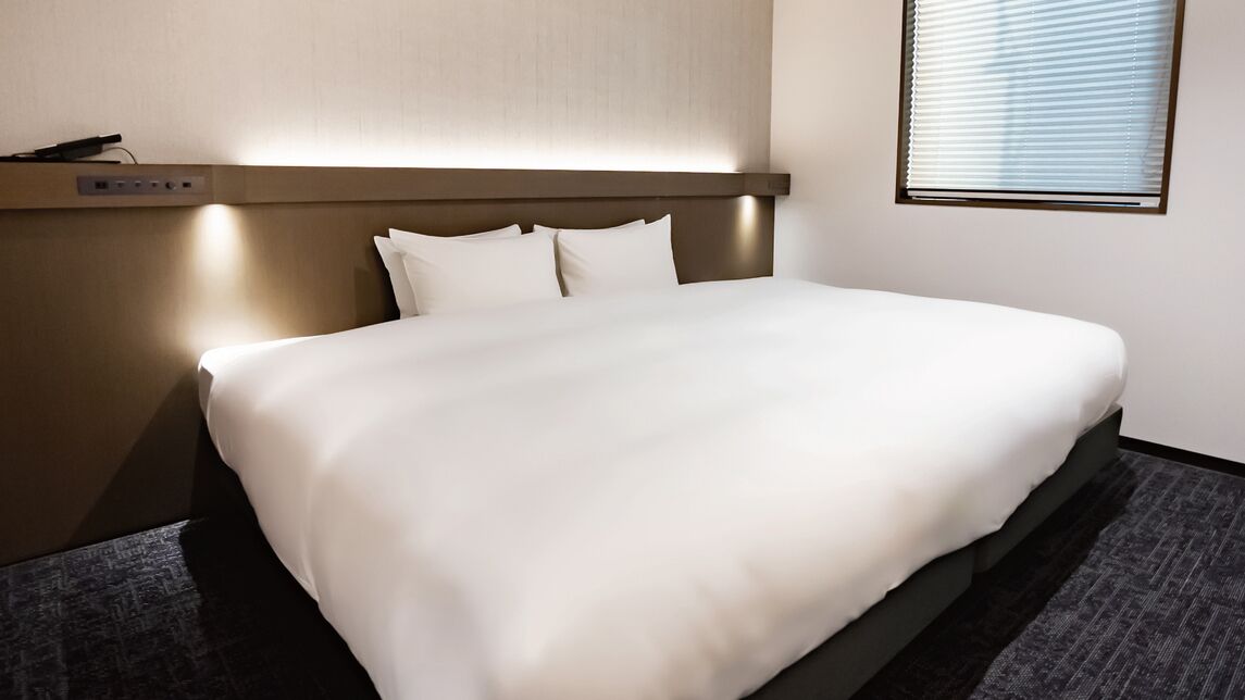 A hotel room with a bed, a headboard with built-in lighting, a power outlet, and a window with blinds.