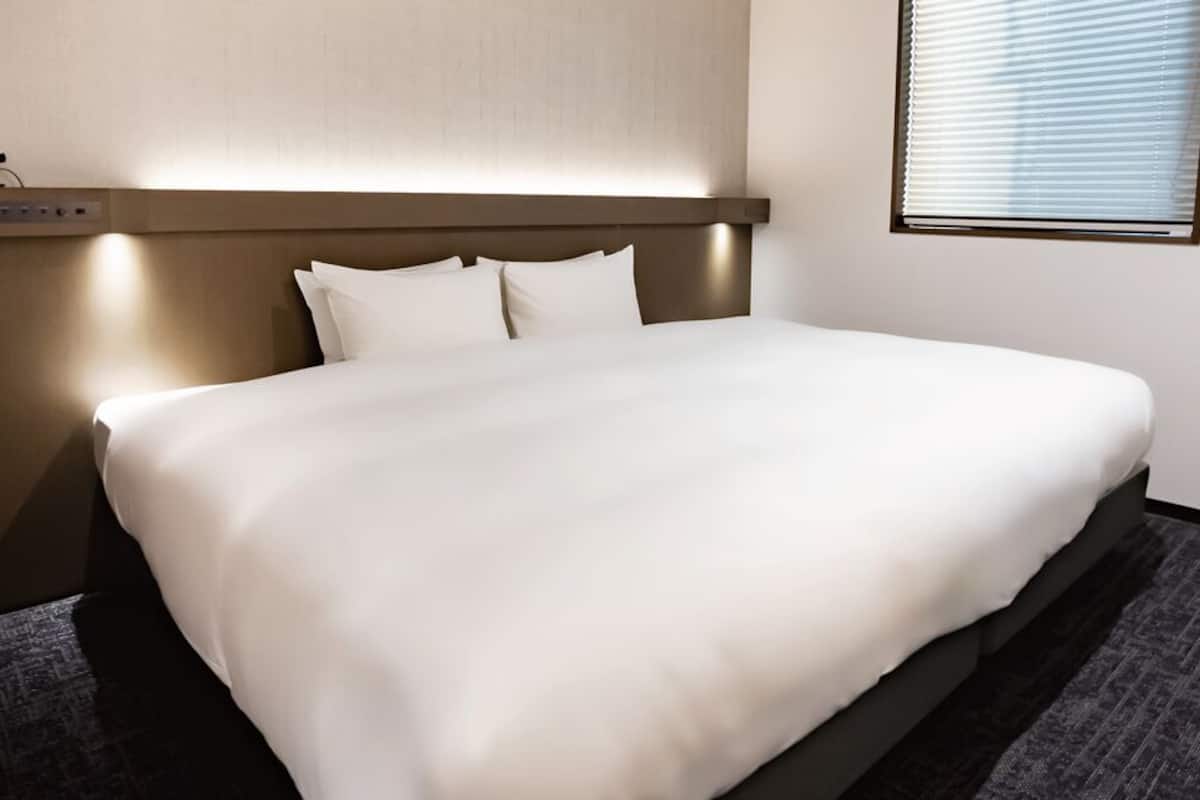 A hotel room with a bed, a headboard with built-in lighting, a power outlet, and a window with blinds.