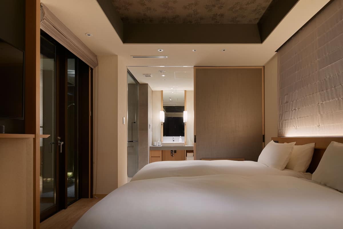 A modern hotel room with a large bed, a walk-in closet, a bathroom with a mirror and sink, and a ceiling with recessed lighting.