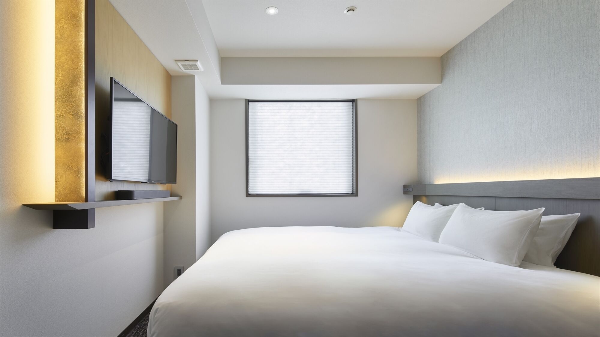 A hotel room with a bed, a television mounted on the wall, a window with blinds, and a ceiling light.