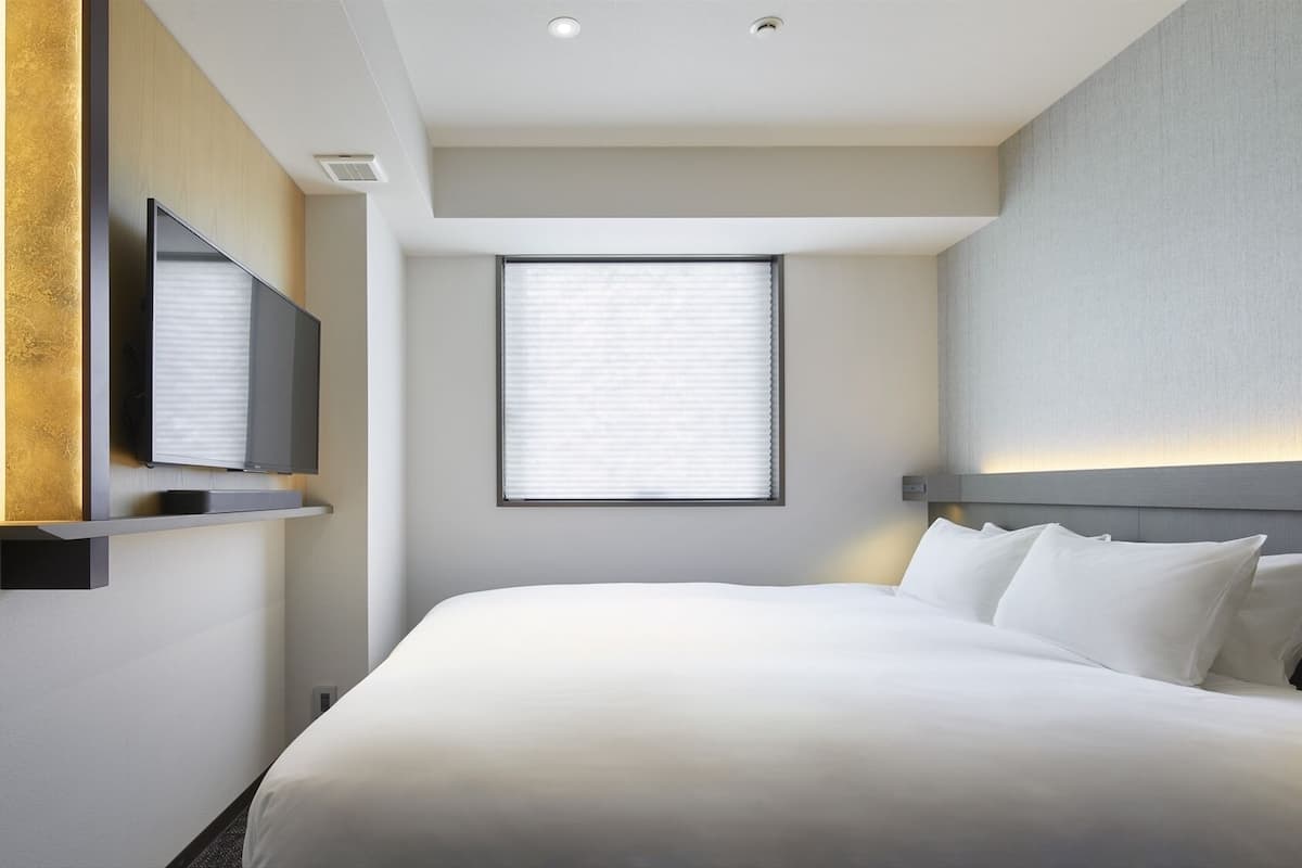 A hotel room with a bed, a television mounted on the wall, a window with blinds, and a ceiling light.