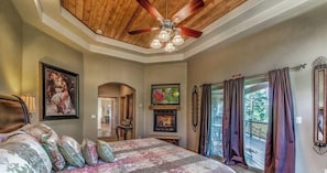 A bedroom with a ceiling fan, a bed with floral bedding, a fireplace, and a view of the outdoors.