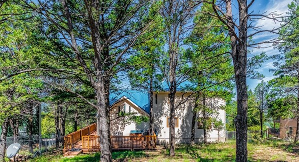 A cabin with a deck and a small porch, surrounded by tall trees and a clear sky.