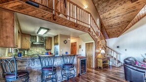 A spacious loft-style living area with a high ceiling and wooden beams, a kitchen with stone accents, and a cozy seating area with a fireplace.