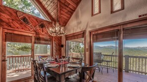 A wooden interior with a dining table set for four, large windows offering a mountain view, and a chandelier.