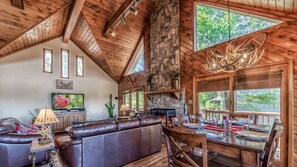 A spacious living area with a stone fireplace, wooden ceiling beams, and a large window overlooking greenery.