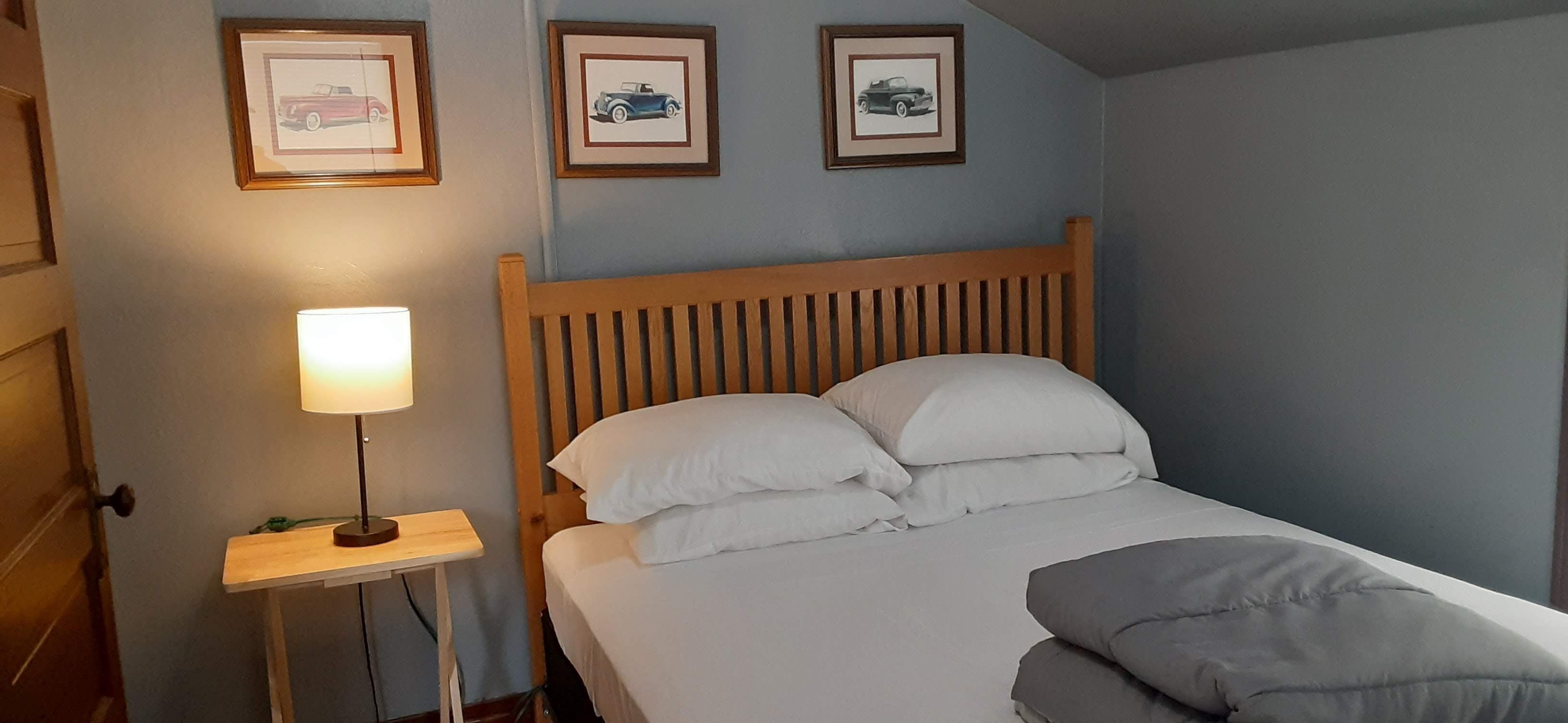 Un lit bien fait, avec du linge de lit blanc et une tête de lit en bois, une table de chevet avec une lampe, et trois tableaux encadrés accrochés au mur.