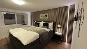Een moderne slaapkamer met een groot bed, een nachtkastje, een wandlamp en een raam met jaloezieën.