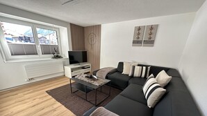 Een moderne woonkamer met een zwarte bank, een salontafel en een televisie. Er is een raam met zonwering, een radiator en een wand van houten panelen.