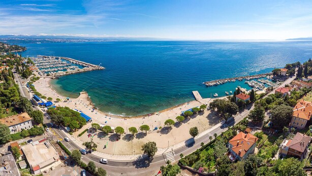 Vista aérea de un pueblo costero con un puerto deportivo, aguas azules cristalinas y una zona de playa con gente y barcos.