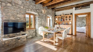 Interior rústico de piedra con una mesa de comedor de madera, sillas, un televisor y una zona de cocina con armarios y vistas al exterior.