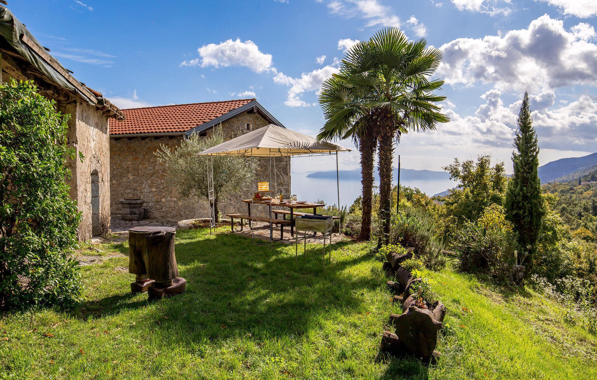 A stone building with a red-tiled roof, a wooden table and benches under a canopy, and a view of the sea.