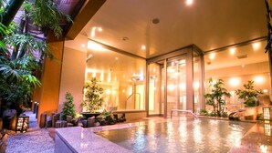 An indoor swimming pool with a glass wall, surrounded by plants and a pebble path. - Dogo Grand Hotel (Matsuyama)