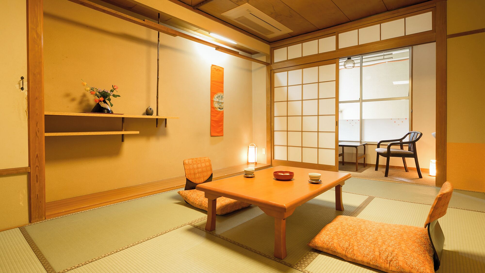 A traditional Japanese room with tatami flooring, a low table, two cushions, a hanging scroll, and a sliding door.