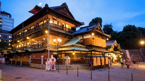 A traditional Japanese building with multiple floors and a tiled roof, featuring a prominent entrance and a small shrine area in front. - Dogo Grand Hotel (Matsuyama)