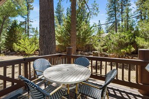 A wooden deck with a round table and four chairs, surrounded by tall trees and a clear sky.