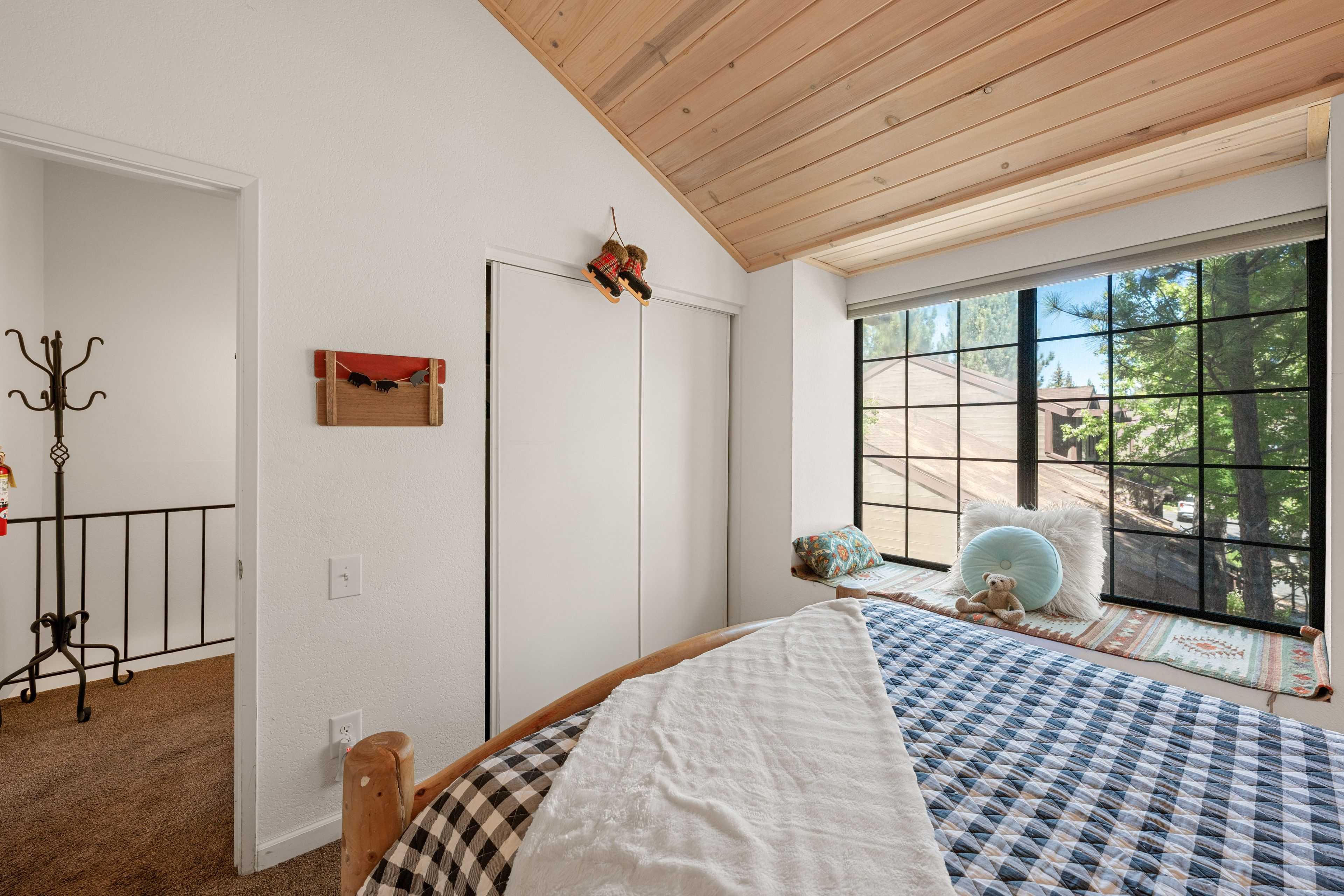 Une chambre avec une grande fenêtre, un lit recouvert d’une couverture à carreaux, un ours en peluche, et une vue sur les arbres à l’extérieur.