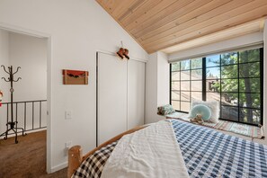 Une chambre avec une grande fenêtre, un lit recouvert d’une couverture à carreaux, un ours en peluche, et une vue sur les arbres à l’extérieur.