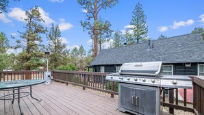 A wooden deck with a grill and a table, surrounded by trees and a building with a dark roof. - Wine Down - Fireplace, Fenced Yard, Deck, Barbecue (Big Bear City)