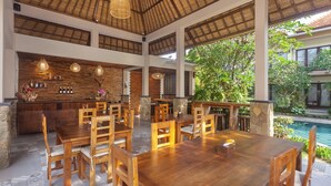 A spacious indoor dining area with wooden tables and chairs, a bar with bottles, and a view of a swimming pool. - The Mudru Resort by GenuineHost (Sukawati)