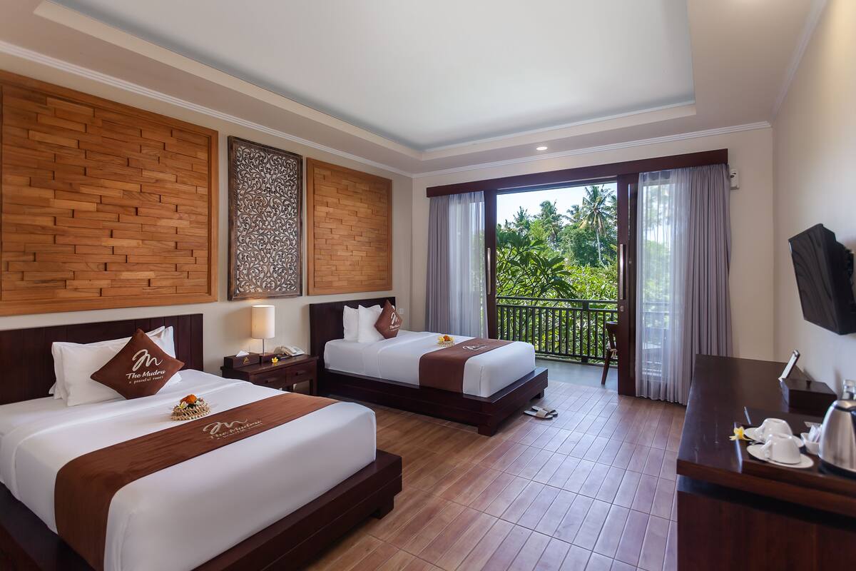 Quarto de hotel com duas camas, uma mesa, uma TV e varanda com vista para área verde.