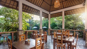 An outdoor patio with wooden tables and chairs, surrounded by lush greenery and a pool. - The Mudru Resort by GenuineHost (Sukawati)