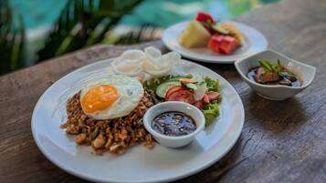Uma refeição com arroz frito, um ovo frito, legumes e um acompanhamento de molho em uma mesa de madeira perto de uma piscina.
