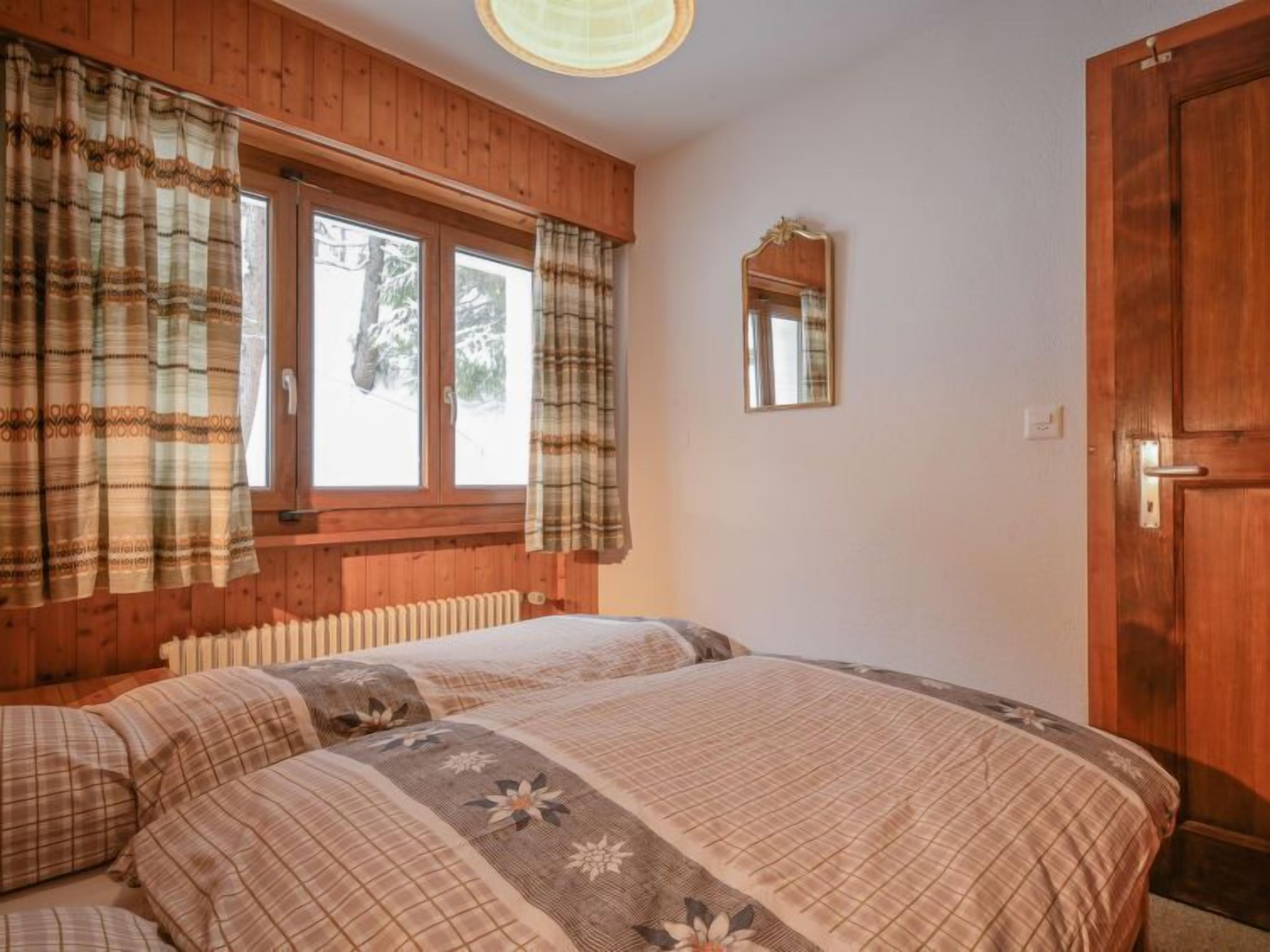 A bedroom with two beds, wooden walls, a window with curtains, a mirror, and a door.