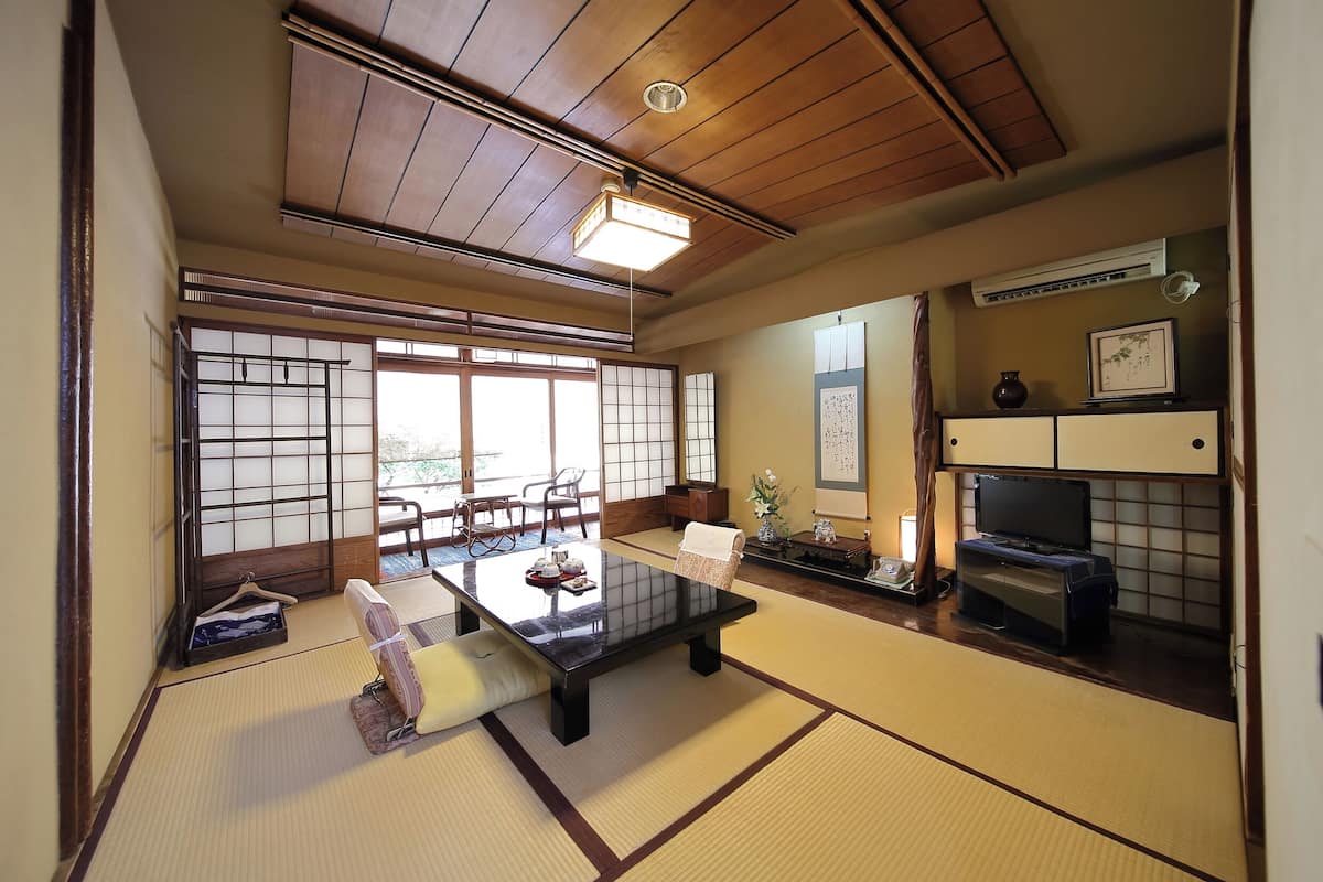 Ein traditioneller japanischer Raum mit Tatami-Boden, einem niedrigen Tisch, einem Fernseher und einer Schiebetür zu einem Essbereich.