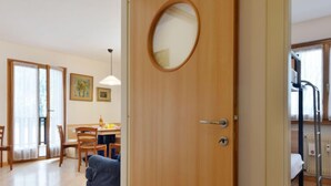 A room with a wooden door, a round mirror, a dining area with chairs and a table, and a bed with a ladder. - Condominio al Parco by Interhome (Canazei)