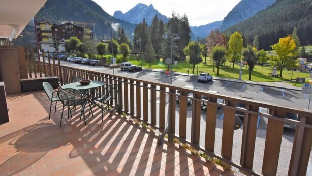 A balcony with a table and chairs, overlooking a street with cars and a park with trees and a playground. - Condominio al Parco by Interhome (Canazei)
