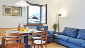 A cozy living room with a dining area, a blue sofa, wooden chairs, a table with plates and a bottle, and a window with a view of a snowy landscape. - Condominio al Parco by Interhome (Canazei)
