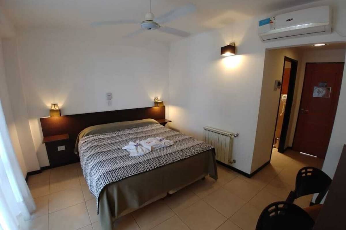 Quarto de hotel com cama, ventoinha no teto, luminárias fixadas na parede, radiador e uma porta.